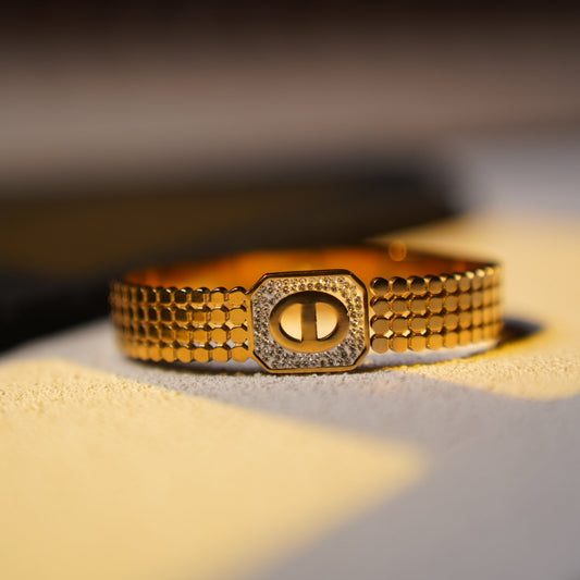 A gold-tone metal bracelet with a textured honeycomb band and a central octagonal clasp embellished with small clear stones, photographed on a light surface with warm directional lighting.