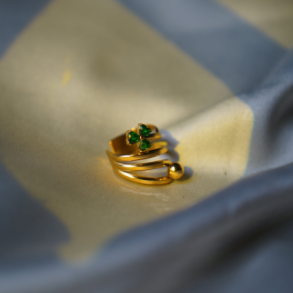 A gold-plated wrap ring with a small green gemstone accent, resting on a draped blue and beige fabric surface.