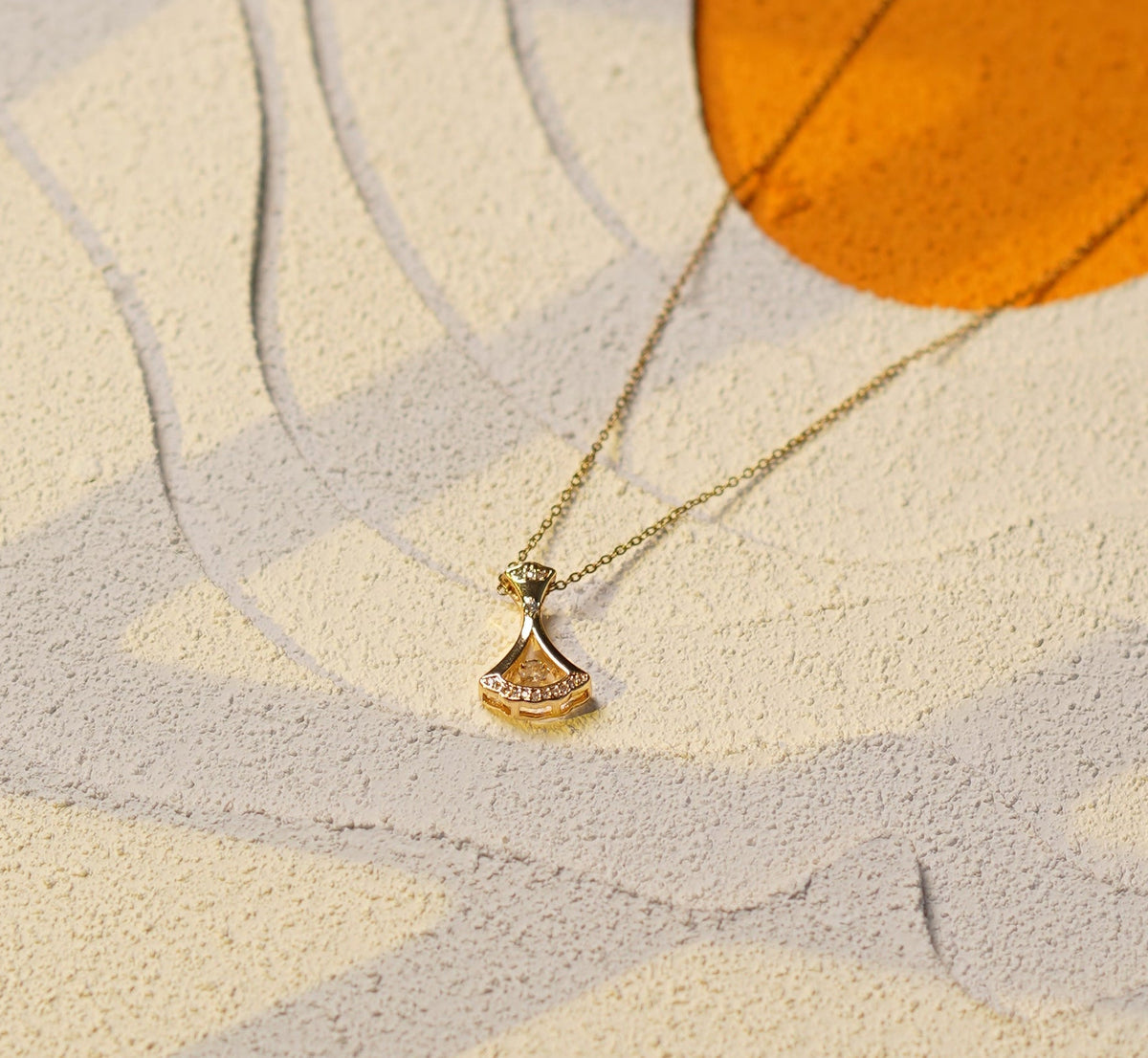 A close-up of a gold necklace with a small round pendant displayed against a textured cream and orange background.