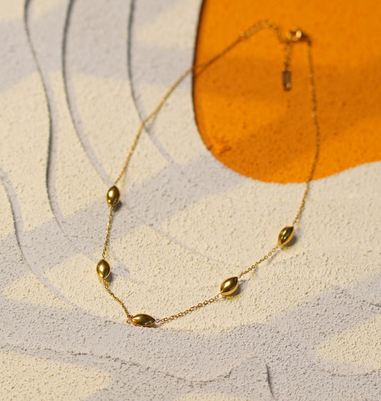 A delicate gold chain bracelet with small bead-like links, laid on a textured abstract surface with cream, gray, and orange shapes.