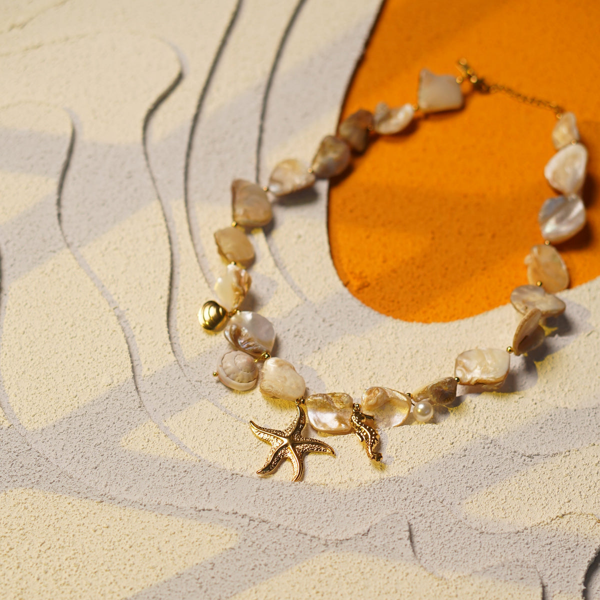A close-up of a beaded bracelet with irregular shell-like pieces in beige, white, and gray tones, accented by a small gold starfish charm and gold-toned metal findings, displayed on a textured abstract surface with gray and orange shapes.