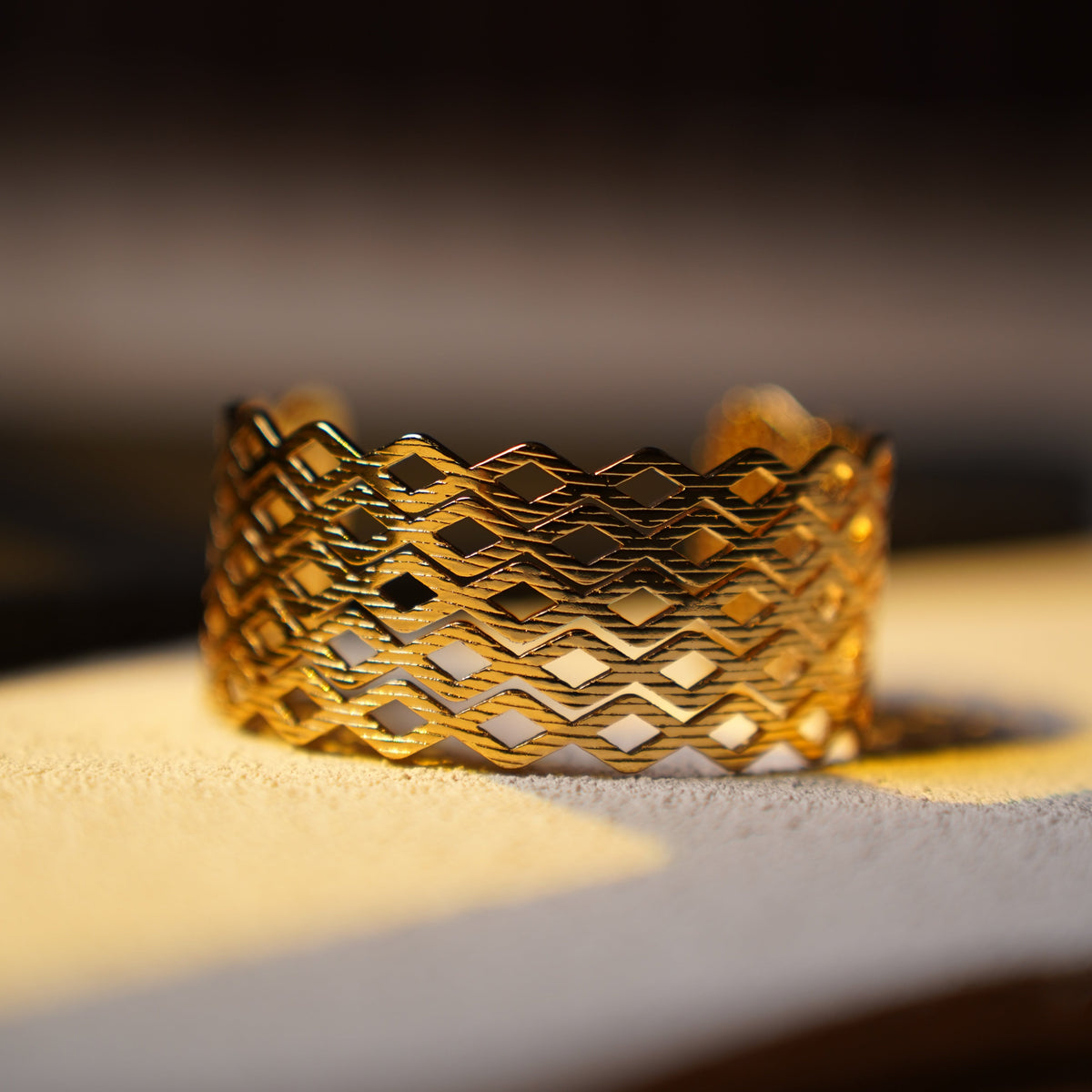 A close-up of a gold-toned cuff bracelet with an intricate openwork geometric pattern, resting on a light-colored surface.