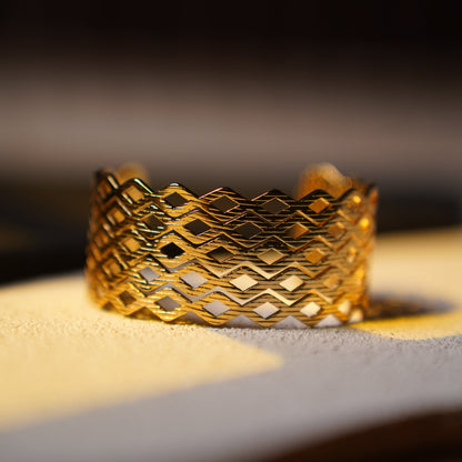 A close-up of a gold-toned cuff bracelet with an intricate openwork geometric pattern, resting on a light-colored surface.