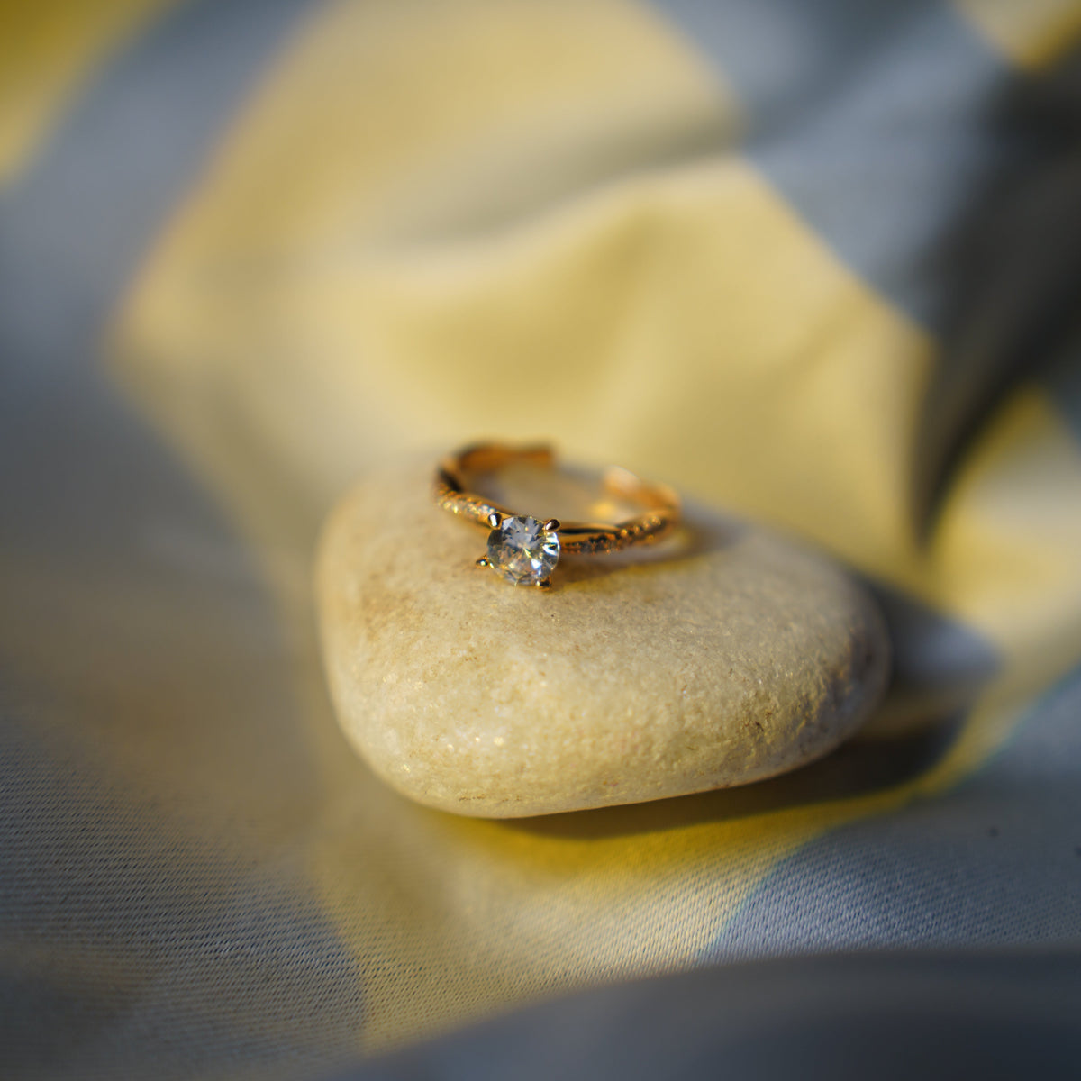 A gold ring with a single clear gemstone resting on a smooth, polished beige stone, set against a soft-focus background of draped yellow and gray fabric.