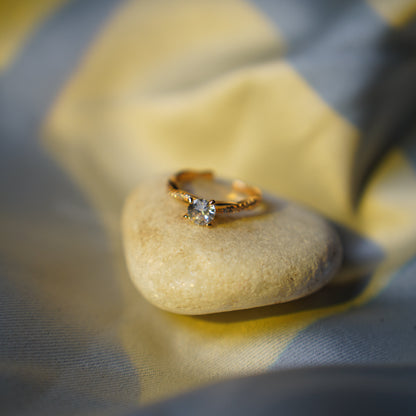 A gold ring with a single clear gemstone resting on a smooth, polished beige stone, set against a soft-focus background of draped yellow and gray fabric.