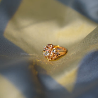 A gold ring with a flower-shaped cluster of clear gemstones, resting on draped gold and blue satin fabric.