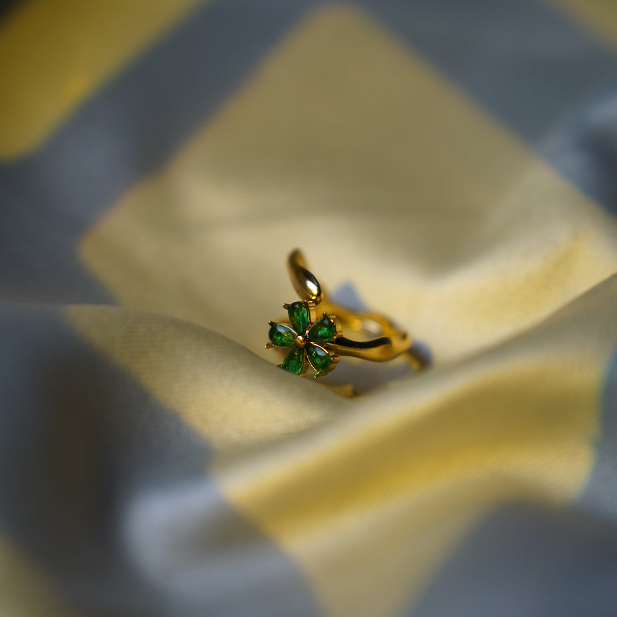 A gold ring with a green four-leaf clover design, resting on a soft, draped fabric background with blue and yellow patterns.