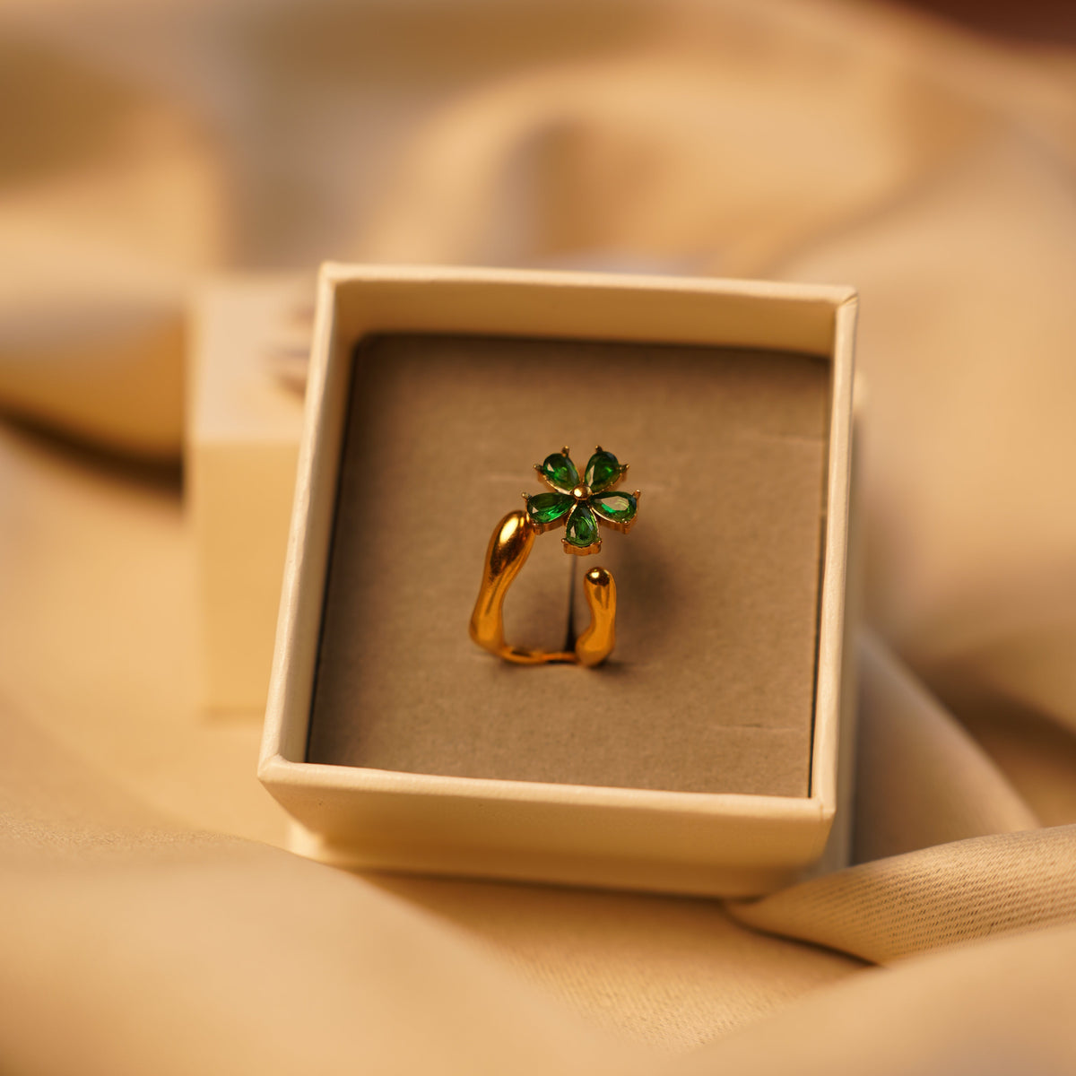 A gold-toned ring with a green four-leaf clover charm, presented in a square cream-colored jewelry box on draped beige fabric.