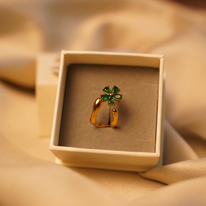 A gold-toned ring with a green four-leaf clover charm, presented in a square cream-colored jewelry box on draped beige fabric.