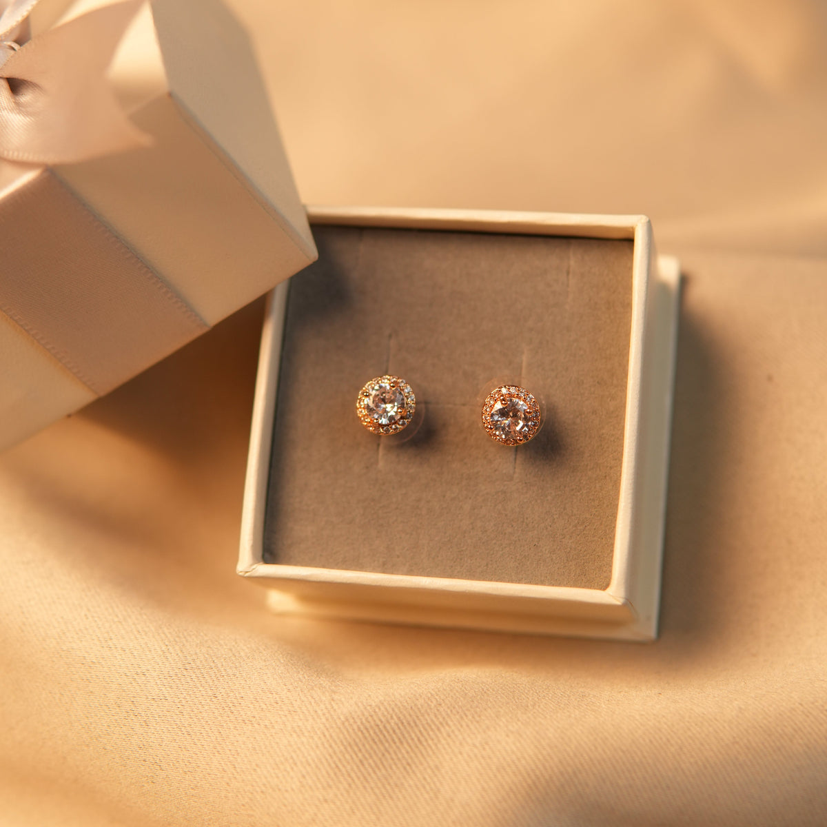 A pair of rose gold stud earrings with clear gemstones, presented in a small cream-colored jewelry box on draped beige fabric.
