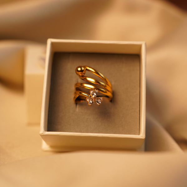 A gold wrap-style ring with a small clear gemstone cluster, displayed inside an open cream-colored jewelry box on draped beige fabric.