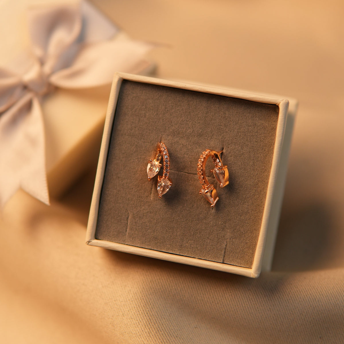 A pair of rose gold hoop earrings with clear crystal accents, presented in a small square gray jewelry box on a warm beige satin fabric background.